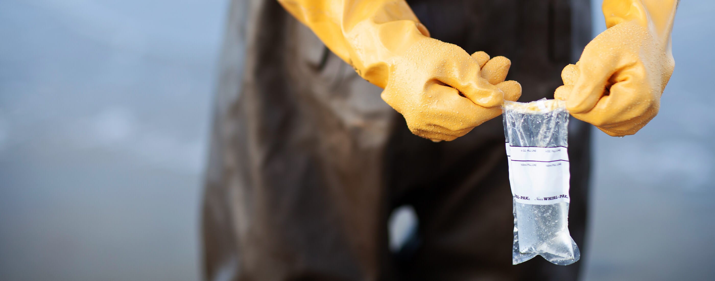 Hands in yellow gloves hold up a sample bag filled with ocean water.