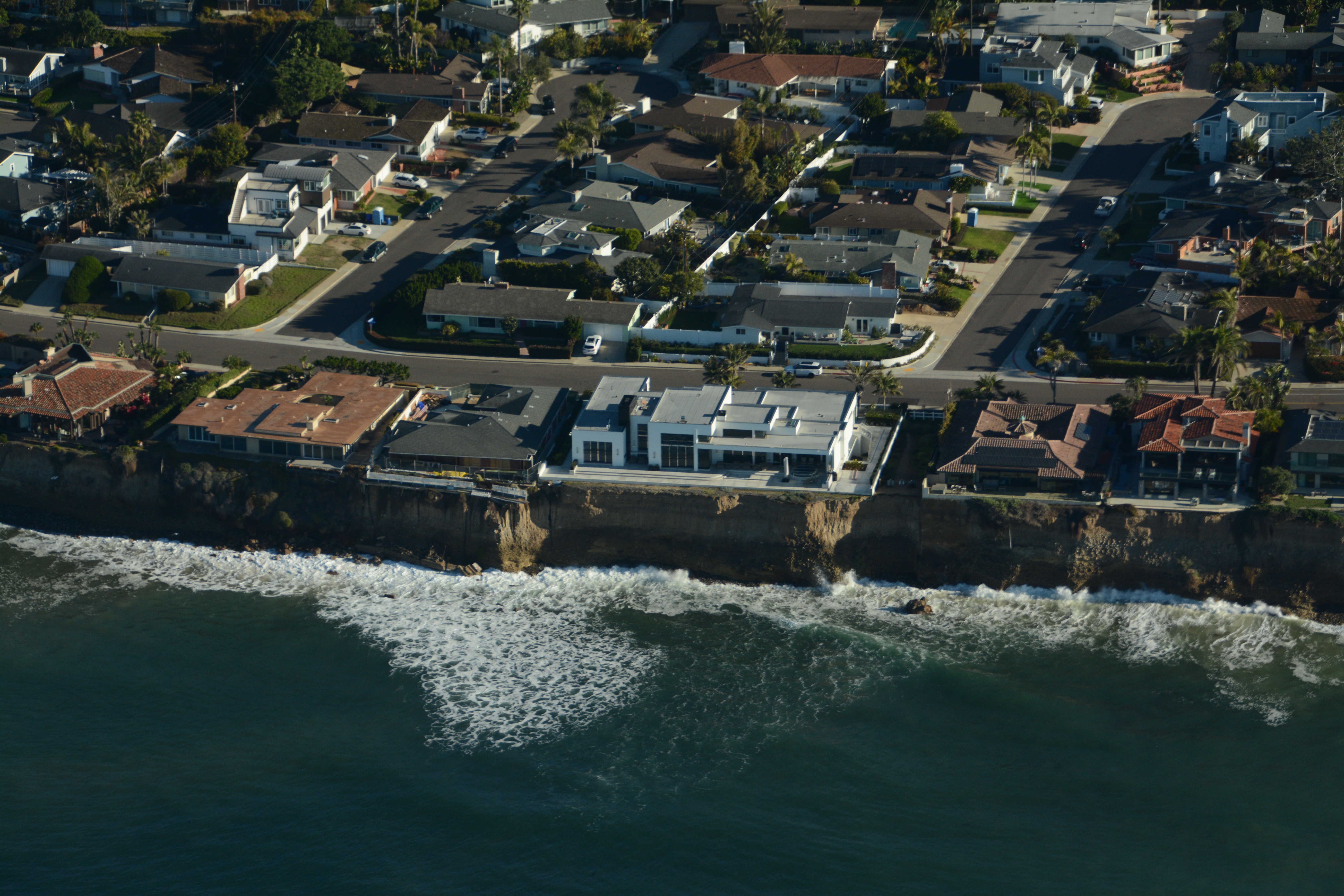 an aerial view from over the ocean looking towards rows of houses on the edge of the ocean bluff. Armored sea walls blocking waves from eroding the cliffs into more beach space.