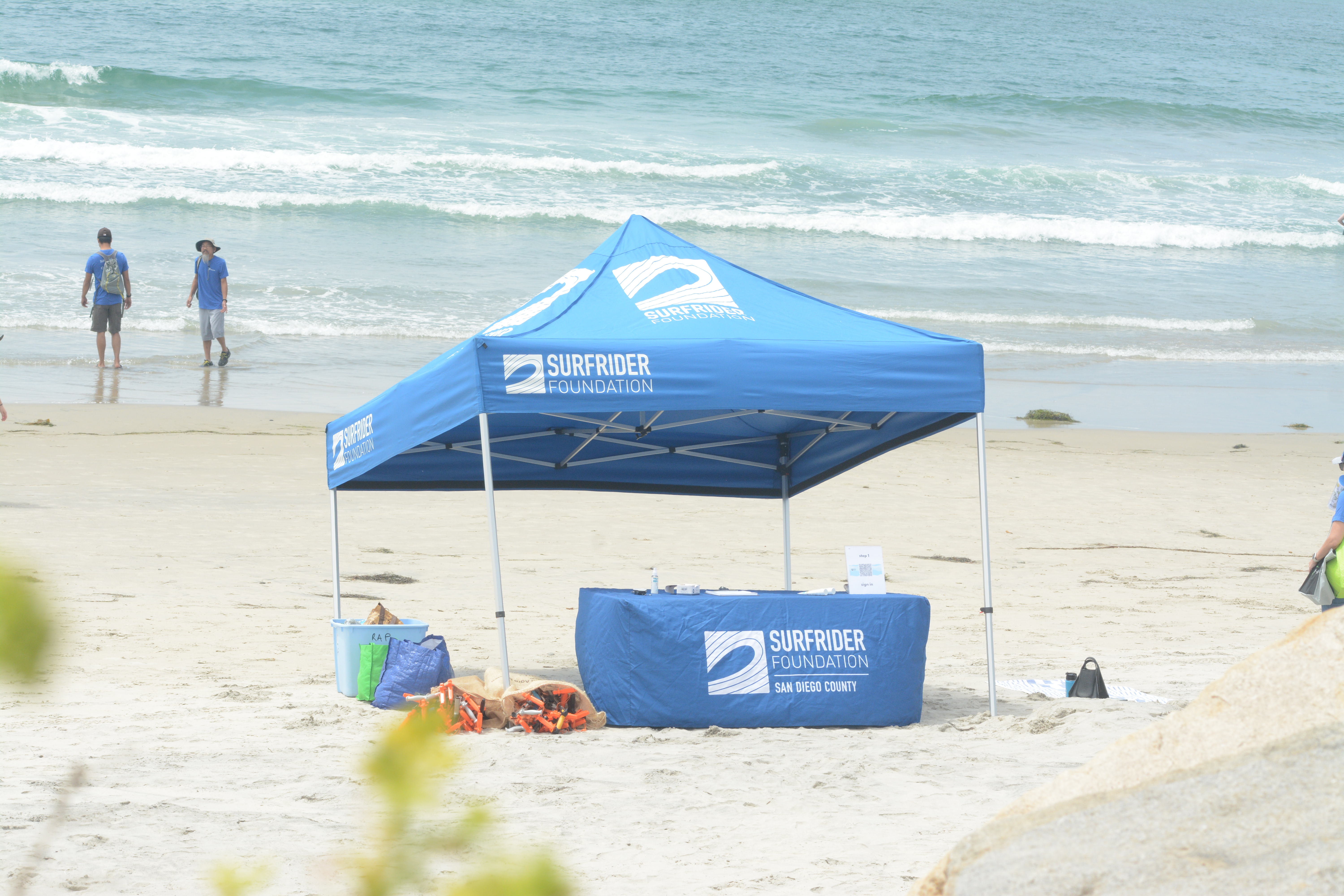 A blue surfrider tent sits on an empty stretch of beach with a blue surfrider table and bags of orange trash grabbers laying on the sand