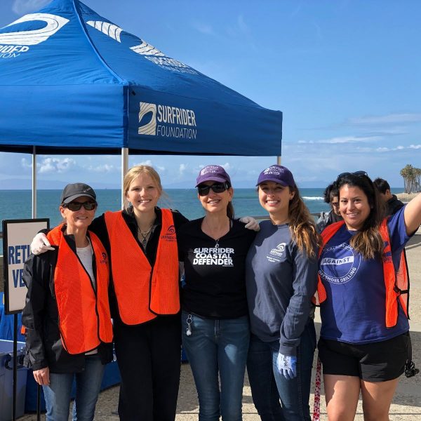 At a Surfrider beach cleanup on the Ventura Promenade. Juli stands in the middle of a group of five awesome volunteers. She's wearing her Surfrider Coastal Defender T-shirt and sunglasses. The Surfrider canopy is behind her, and behind that, a beautiful day on the Pacific Ocean.