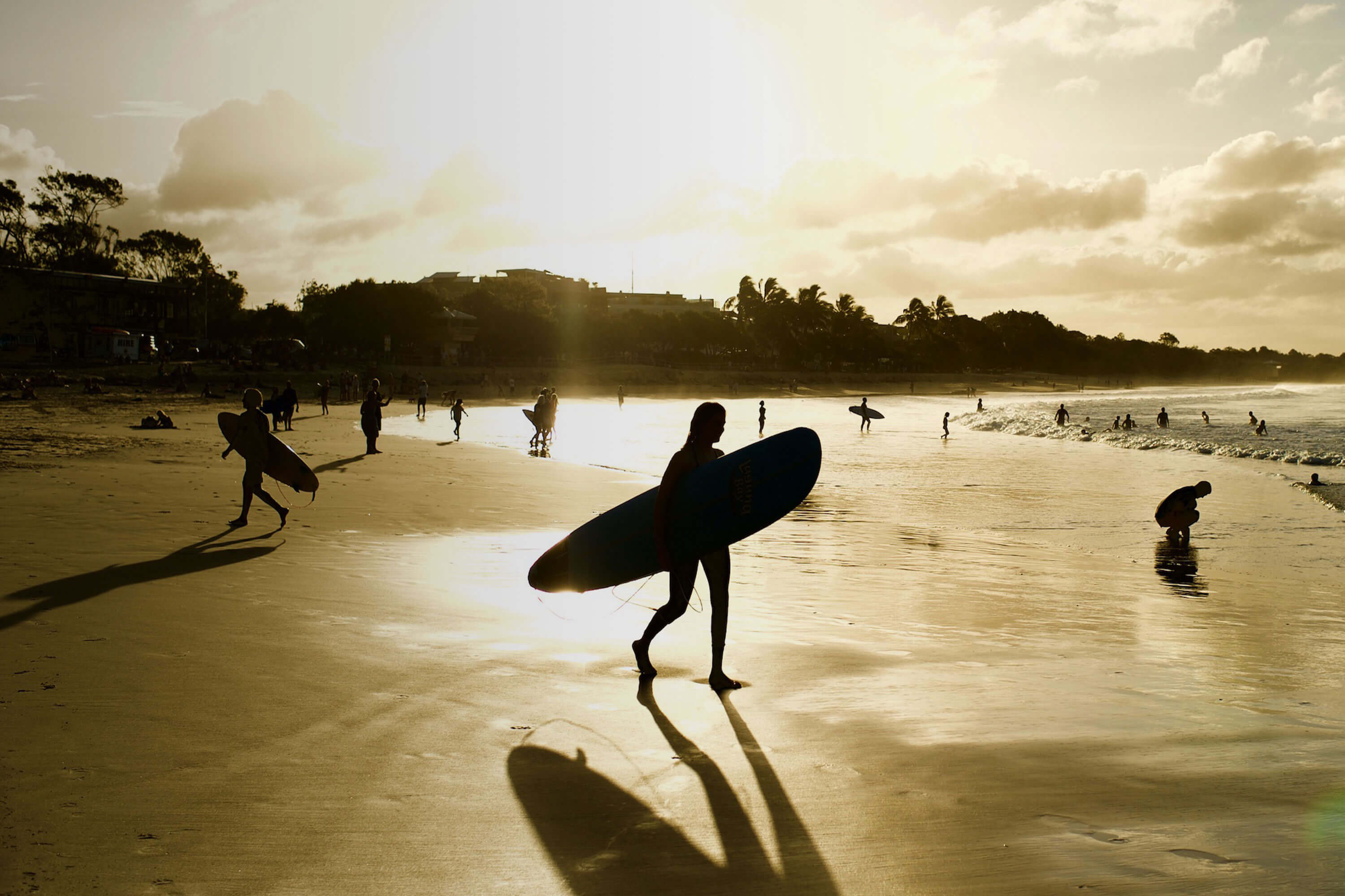Surfers walk along a beach during the golden hour of sunset.