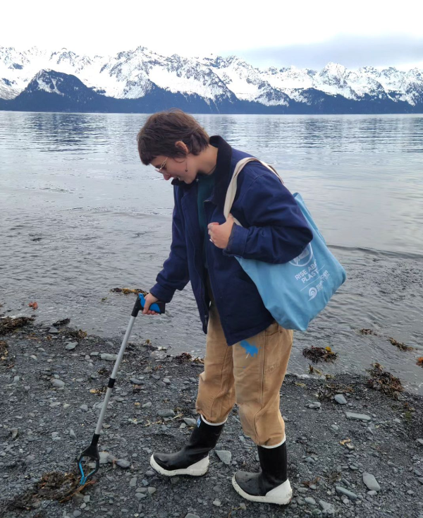 A boot clad volunteer picks up trash on a pebble beach with snowy peaks in the  background