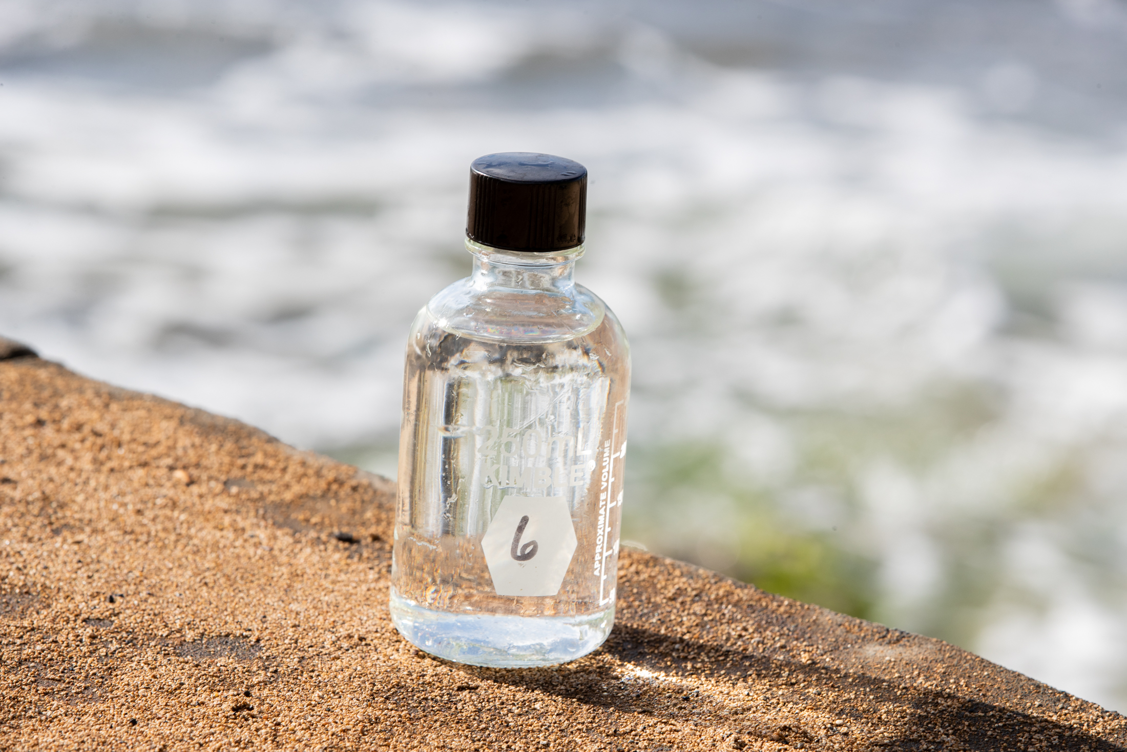 Water sample bottle sitting full infront an out of focus beach