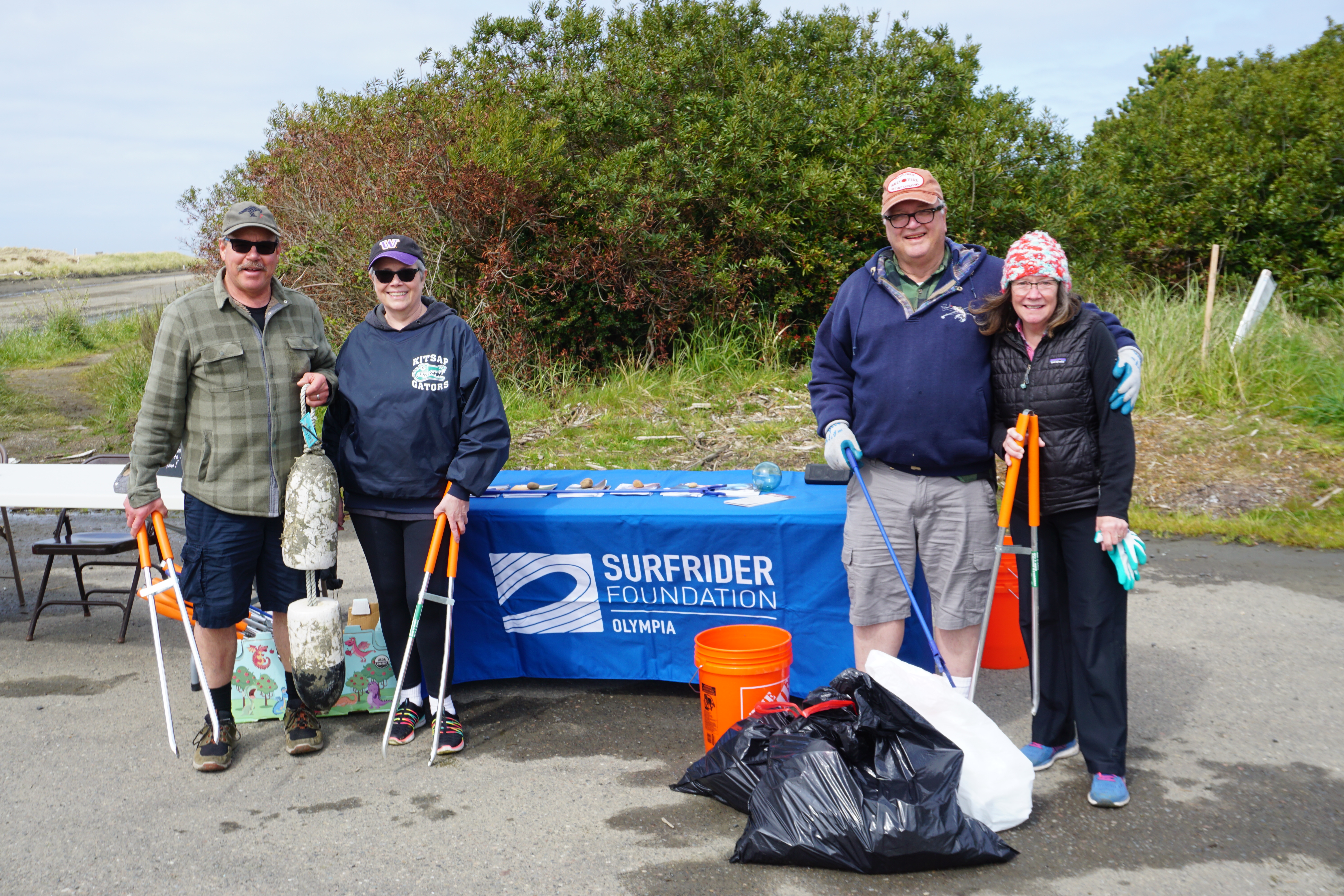 4 volunteers pose by our cleanup table with trash pickers and bags of debris 