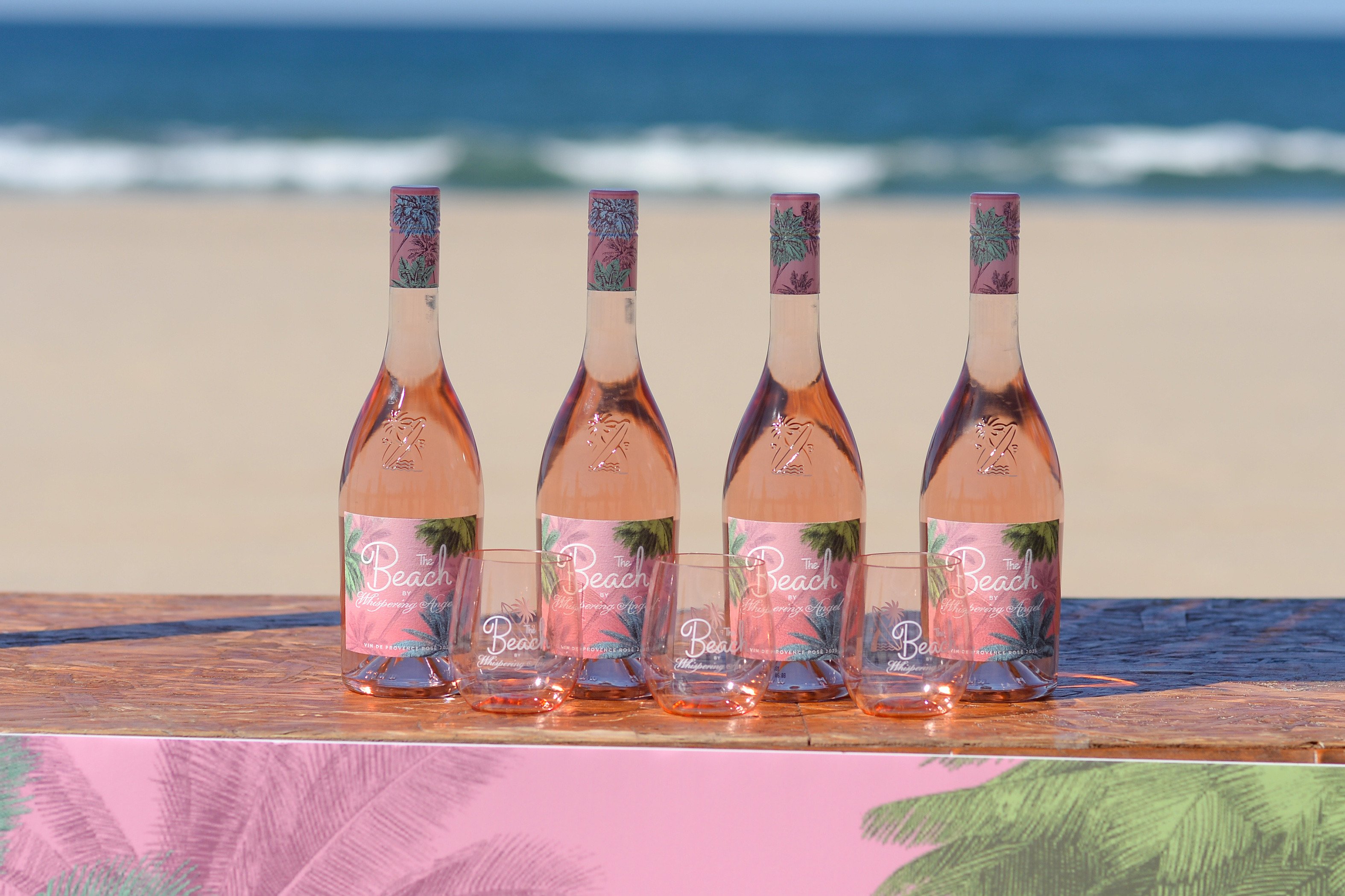 Four bottles of The Beach rosé with four glasses in front of the bottles at the beach