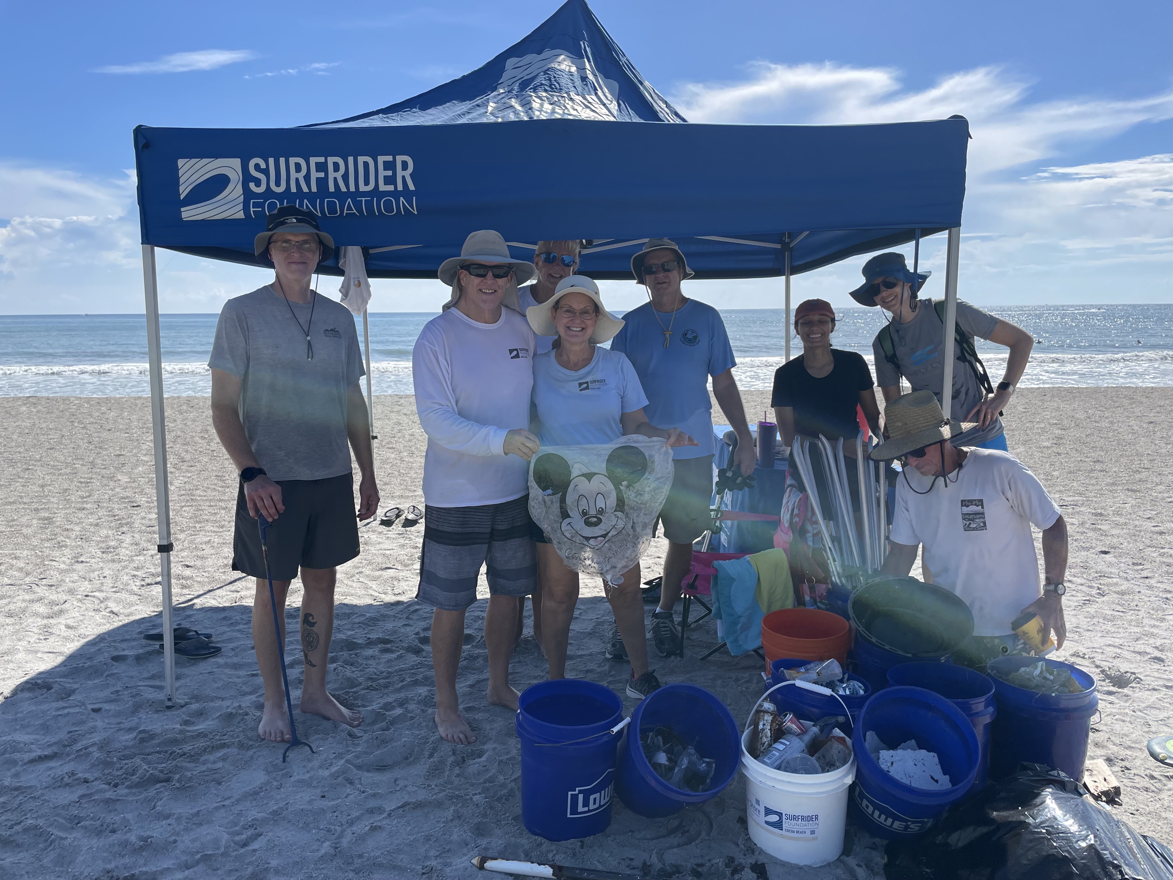Group of volunteers on the beach with beach cleanup tools, standing under a blue tent with the Surfrider logo on it