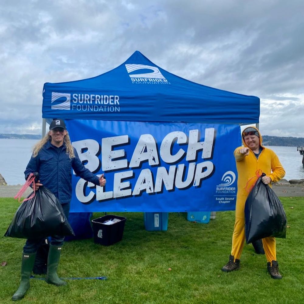 Two volunteers posing with full garbage bags in front of a blue canopy and beach cleanup sign 