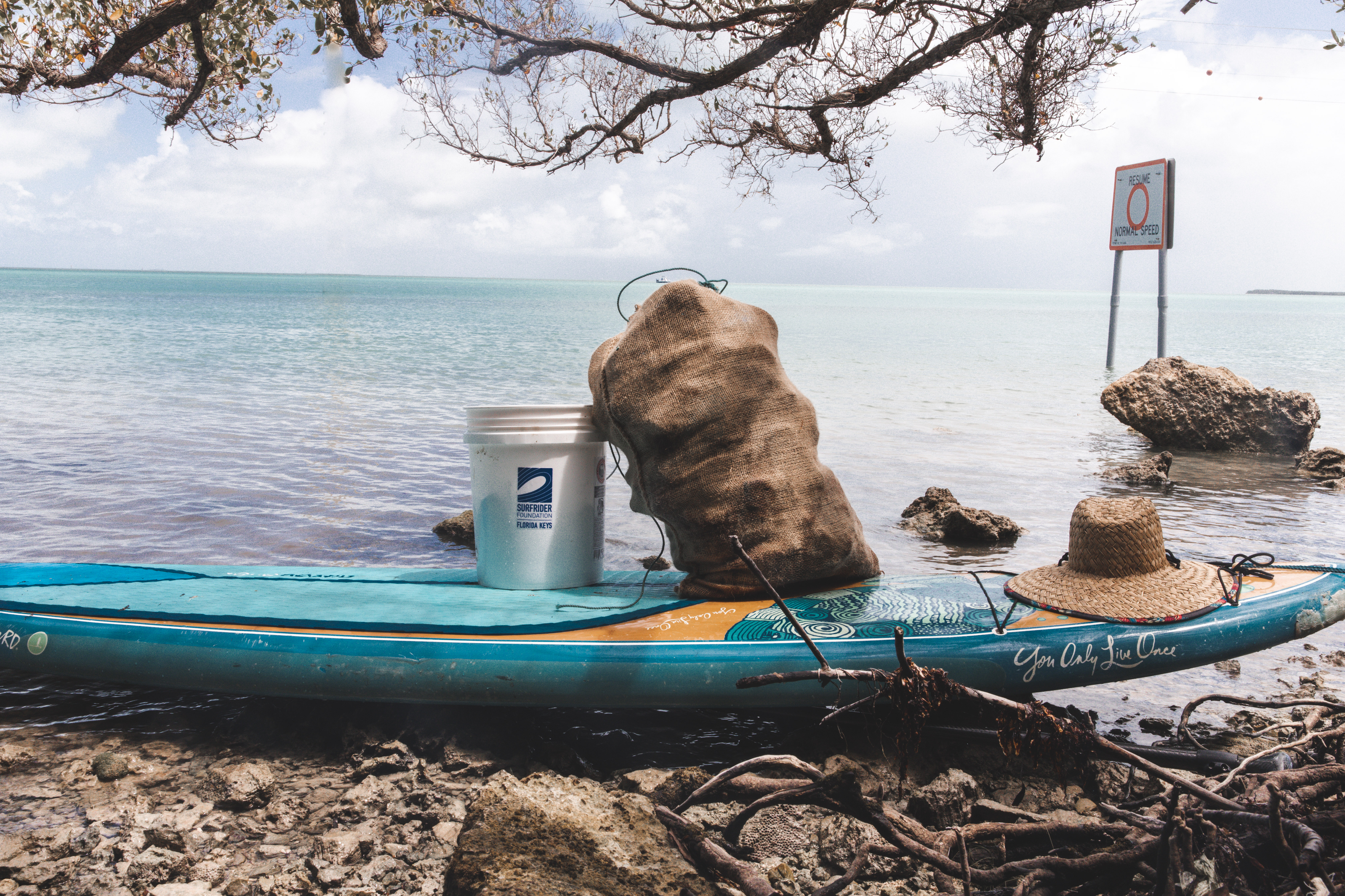 A bag of trash, a bucket, and bag of trash sit on a surf board near the water