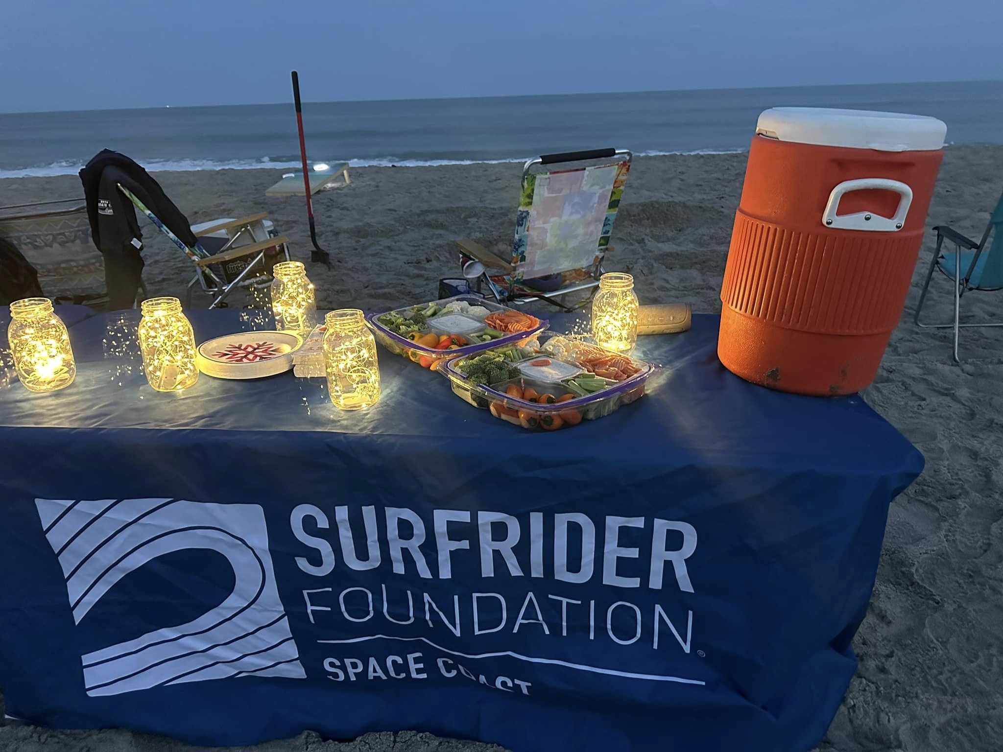 A table on the beach with a Surfrider-labeled tablecloth, topped with a water jug, lights, and veggie trays