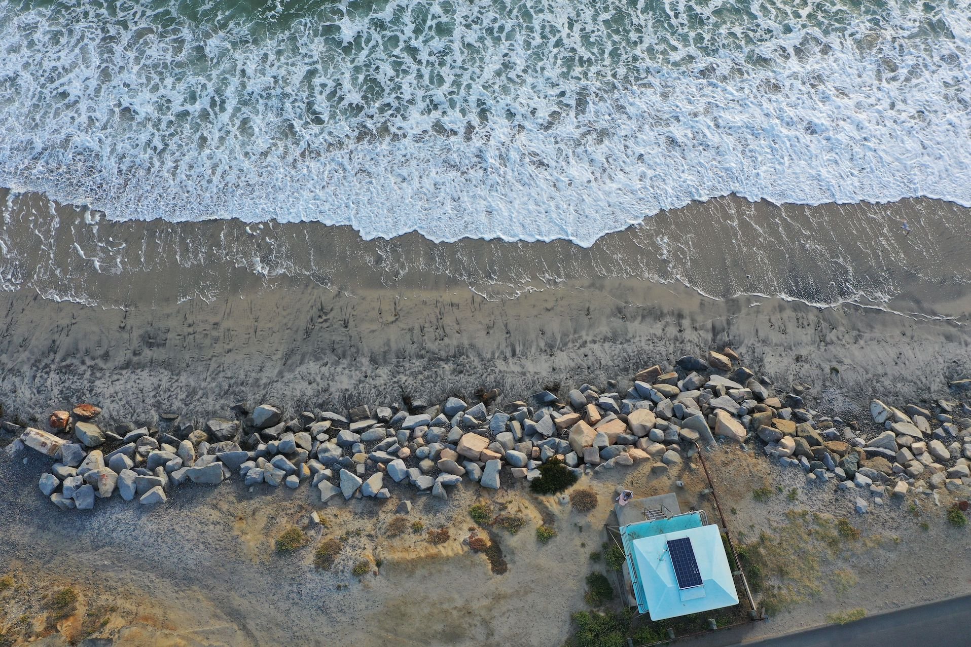 overhead drone image of a narrow, boulder-lined beach in front of Southbound Carlsbad Blvd.