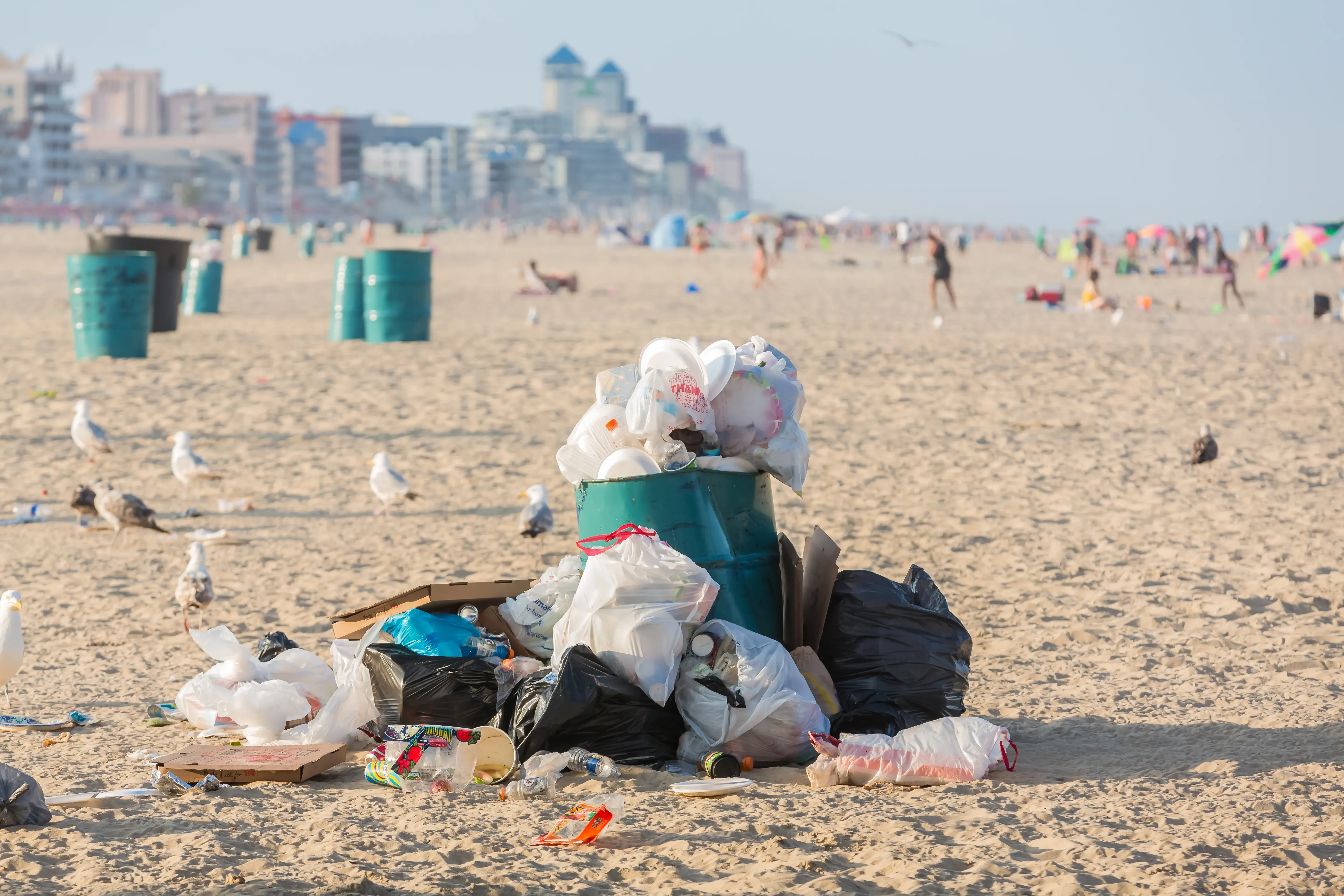 Trash piled around a waste bin on a sandy beach.
